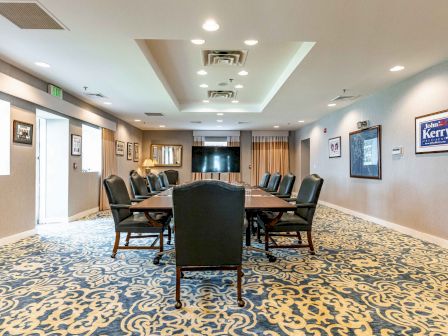 A conference room with a long table, multiple chairs, patterned carpet, framed pictures on walls, and a large TV screen at the far end.