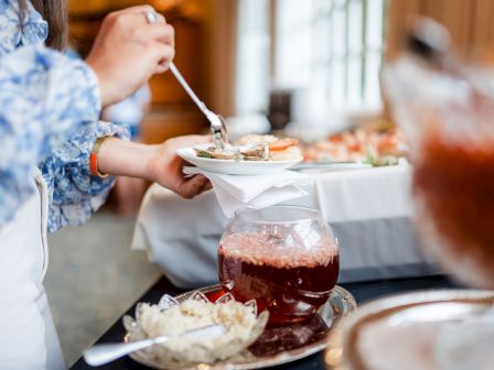 A person is serving food onto a plate at a buffet table with various dishes, including a bowl of red sauce and a bowl of chopped onions.