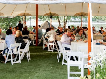 A group of people are seated at round tables under a tent, participating in an outdoor event or gathering by a body of water.