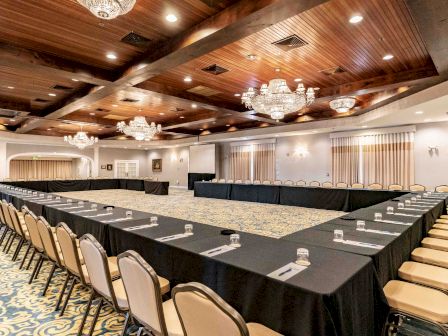 A well-lit conference room with U-shaped table setup, adorned with chandeliers, beige chairs, and notepads with pens on the tables, ready for attendees.