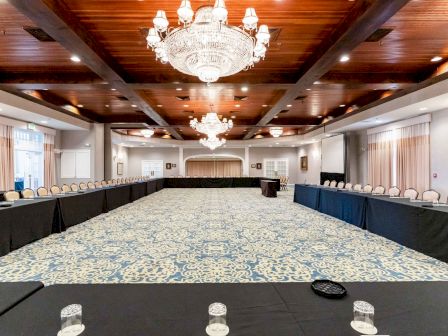 A large conference room with chandeliers, U-shaped table arrangement, black tablecloths, chairs, and a patterned carpet is shown in the image.