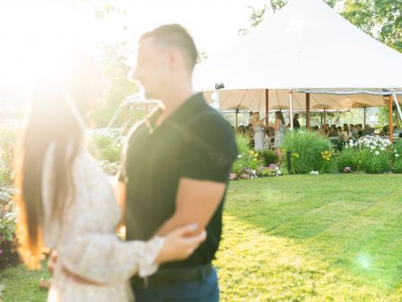 A couple stands facing each other outdoors, with a blurred background of a large white tent and guests in a garden setting.