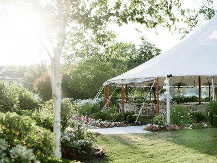 An outdoor garden venue with lush greenery and flowers features a white tent canopy covering tables and chairs, creating a serene event space.