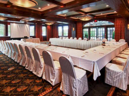 A conference room setup with U-shaped table arrangement, white chair covers, projector, screen, and elegant lighting, ready for a meeting or event.