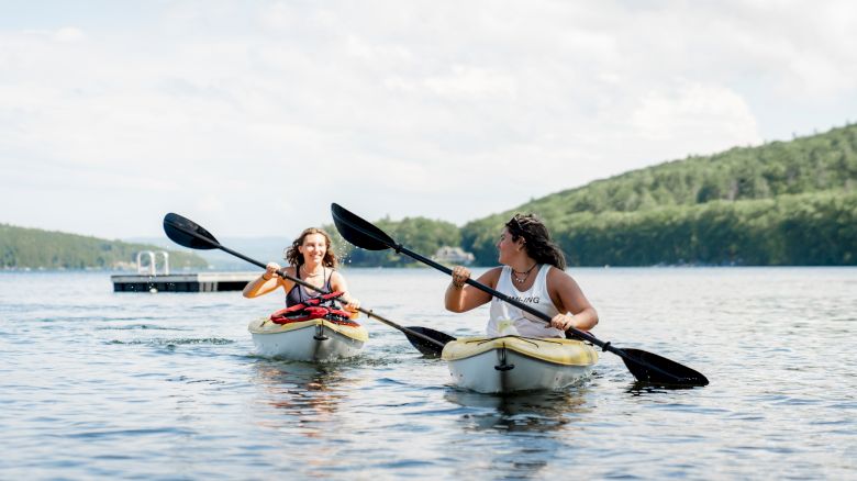 Two people are kayaking on a calm body of water with a dock in the background, surrounded by lush green hills under a partly cloudy sky.