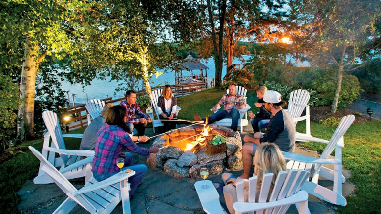 A group of people sits in a circle of Adirondack chairs around a fire pit, surrounded by trees, with a body of water in the background ending the sentence.