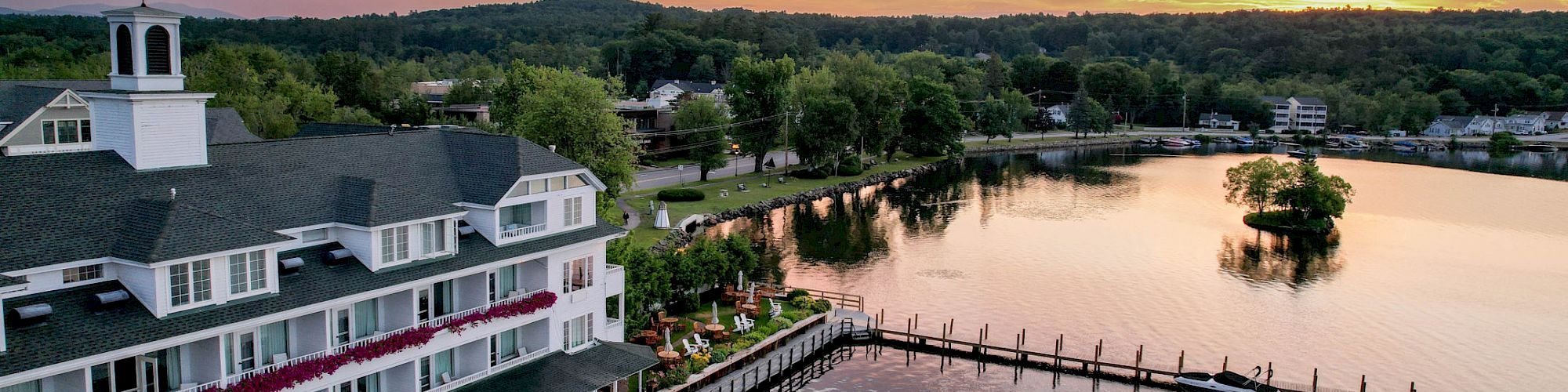 A beautiful lakeside building with a dock extending into the water during a picturesque sunset, surrounded by trees and a small island.