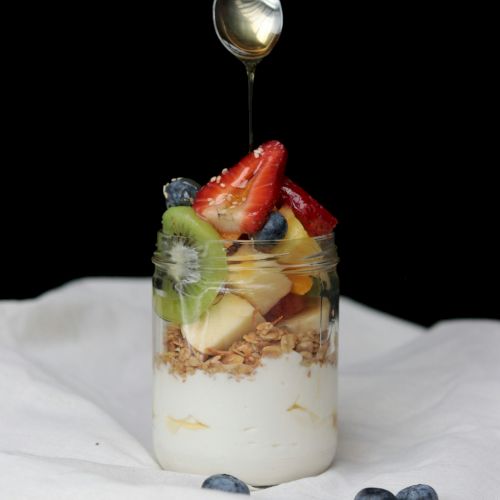 A jar of yogurt with granola, kiwis, strawberries, and blueberries, drizzled with honey from a spoon above, placed on a white cloth.