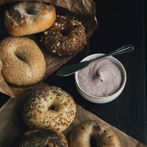 An assortment of bagels arranged on a paper with a bowl of cream cheese and a knife beside them, on a dark surface.