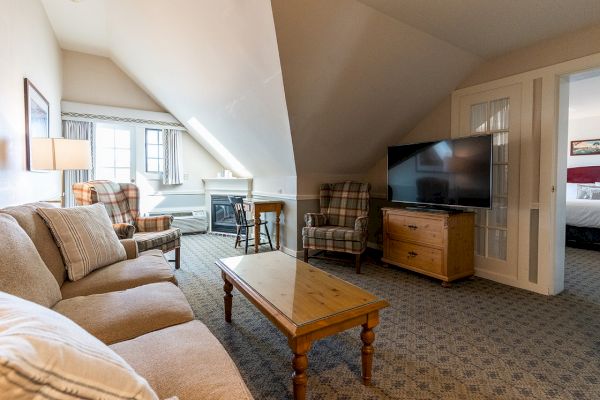 The image shows a cozy living room with a sofa, armchairs, coffee table, large TV, and a glimpse of a bedroom in the background, all in soft lighting.