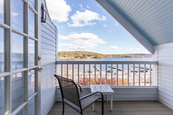 A balcony with a chair and table overlooks water and boats under a clear sky, with white railing and siding completing the serene view.