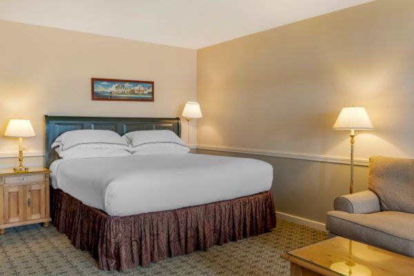 The image shows a hotel room with a king-sized bed, two nightstands with lamps, a beige sofa, a glass-top coffee table, and a framed picture above the bed.