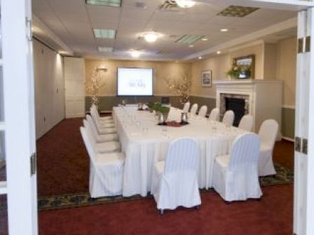 The image shows a conference room with a U-shaped table arrangement, white covered chairs, a projector screen, and a fireplace with a mirror above it.