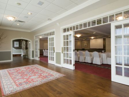 The image shows an elegant room with hardwood floors, a red patterned rug, and white-framed French doors opening to a dining or meeting area.