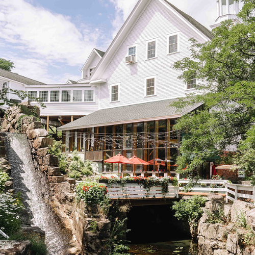 A charming building adorned with white railings and surrounded by lush greenery, featuring a flowing waterfall and an outdoor dining area with red umbrellas.