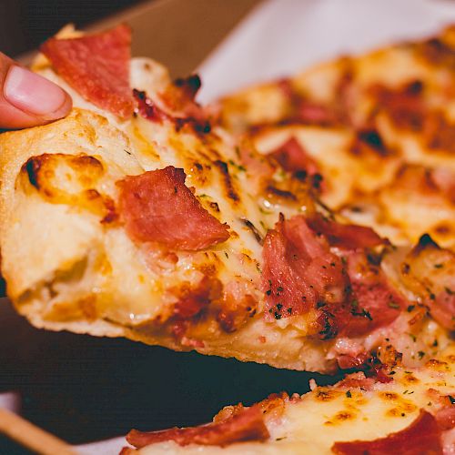 A hand is holding a slice of pizza topped with cheese and chunks of meat from a pizza box.