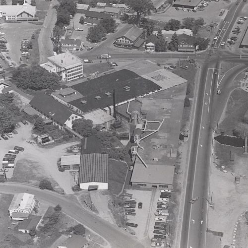 An aerial view of a small town with buildings, roads, vehicles, and some waterfront areas, showing a mix of residential and commercial structures.