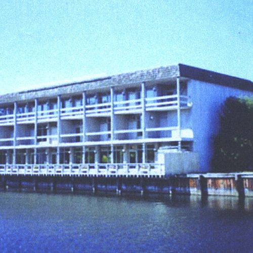 The image shows a multi-story building with balconies, situated beside a body of water, likely a river or lake, surrounded by some trees and foliage.