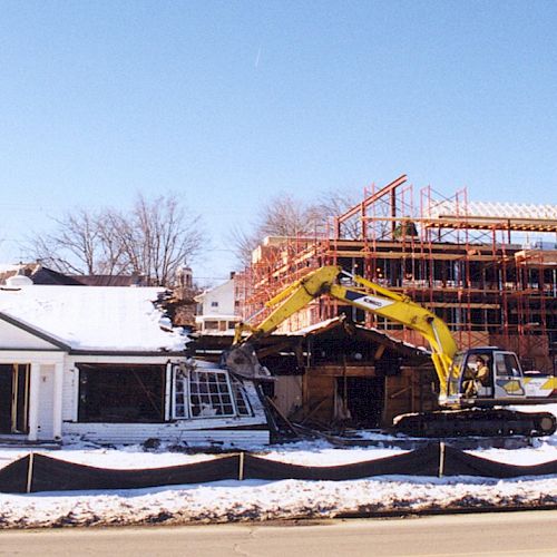 An excavator is demolishing a small building with a larger construction project visible in the background on a snowy day.