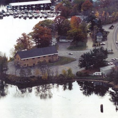 An aerial view of a lakeside area with a large building, marina, boats, and a winding road through a forested region, possibly in autumn.