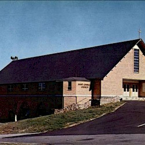 The image shows a church building with a slanted roof, set near a body of water, with a parking lot in the foreground and trees in the background.