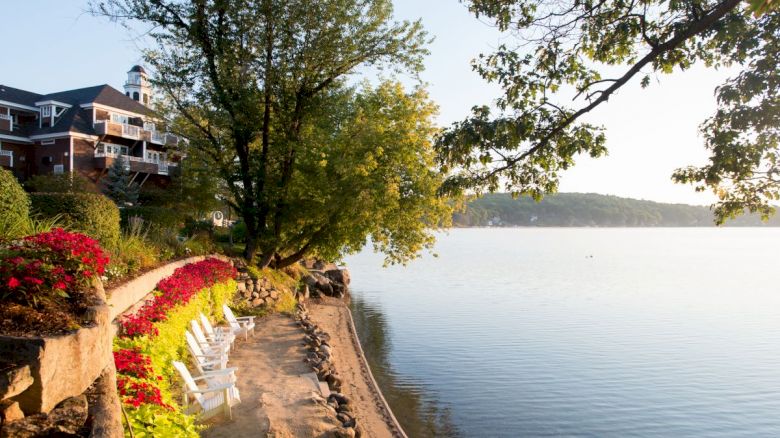 A lake with a sandy shore, white chairs, a tree, a building with balconies, and flowering plants under a clear sky at sunset.