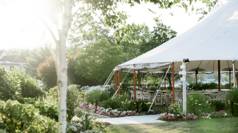 A scenic garden with a white tented event area surrounded by greenery and colorful flowers, trees, and a pathway leading to the tent.