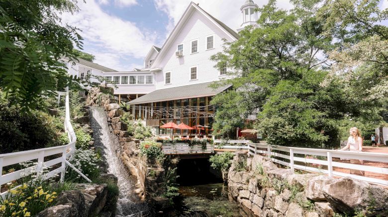 The image shows a picturesque building with a white facade surrounded by lush greenery, a waterfall, outdoor seating, and a small bridge.