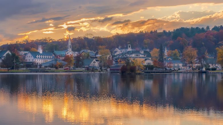 A picturesque town by a lake during sunset, with autumn foliage, reflections on the water, and a serene, peaceful atmosphere ending the sentence.