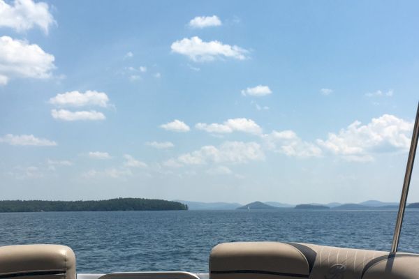 The image shows a serene view from the back of a boat, looking out over a calm lake with distant islands and a partly cloudy sky.