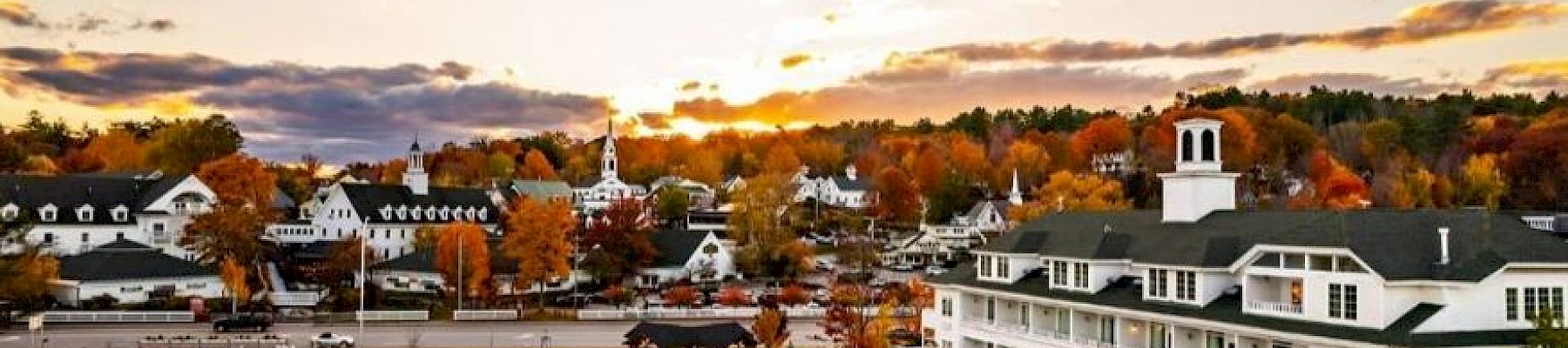 A scenic lakeside town with white buildings, autumn foliage, and a sunset sky reflected in the water.