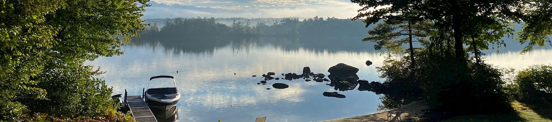 A serene lakeside scene with a small boat docked, calm water, trees on both sides, rocks in the water, and a partly cloudy sky.