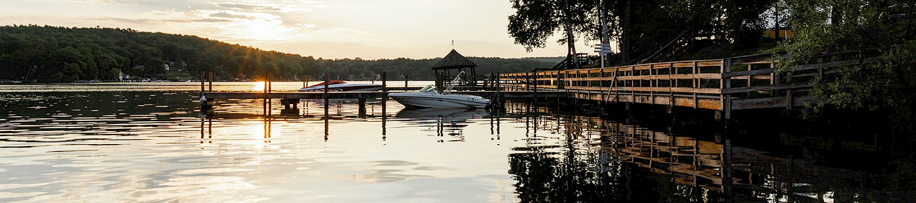 A tranquil lakeside scene during sunset, featuring a wooden pier, docked boats, and trees reflected in the calm water under a partially cloudy sky.