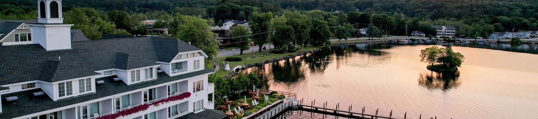 An elegant lakeside building at sunset with a dock extending into the calm waters, surrounded by lush greenery, under a partly cloudy sky.