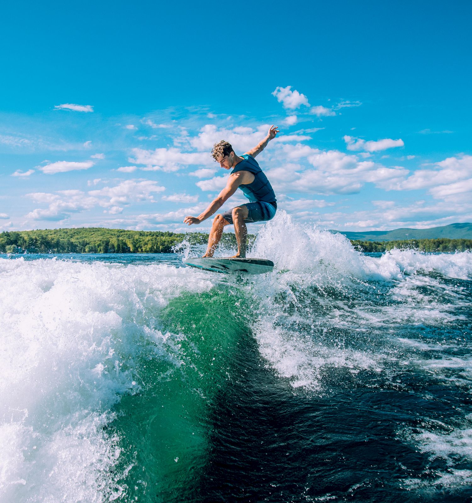 A person is wakesurfing on a large wake in a body of water, surrounded by a scenic landscape with trees and mountains under a bright blue sky.