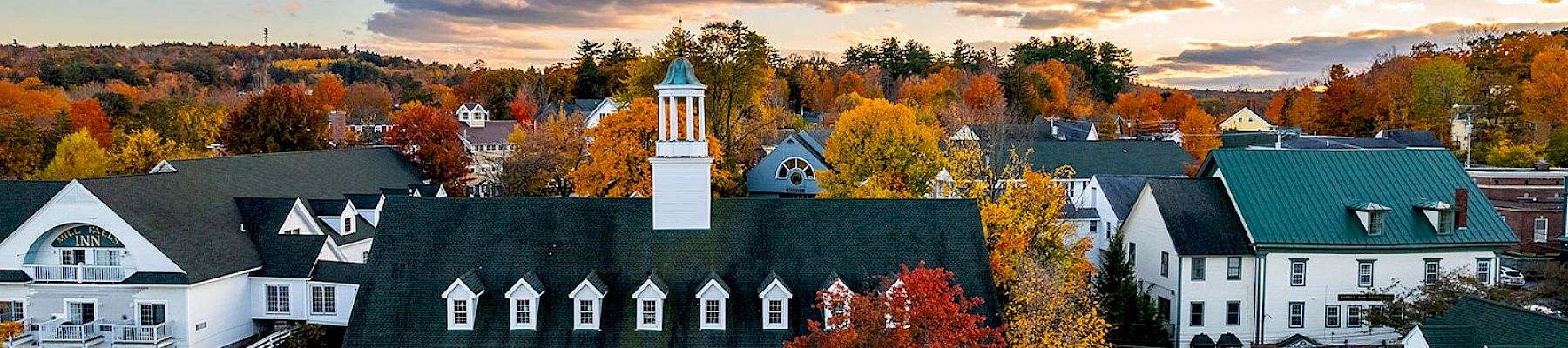 A picturesque town with white buildings, green roofs, and a central clock tower surrounded by vibrant autumn foliage under a partly cloudy sky.