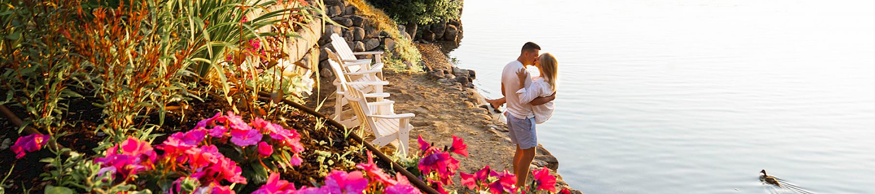 A couple embraces beside a lake surrounded by vibrant flowers and greenery, with a scenic house and a tranquil day on the water in the background.