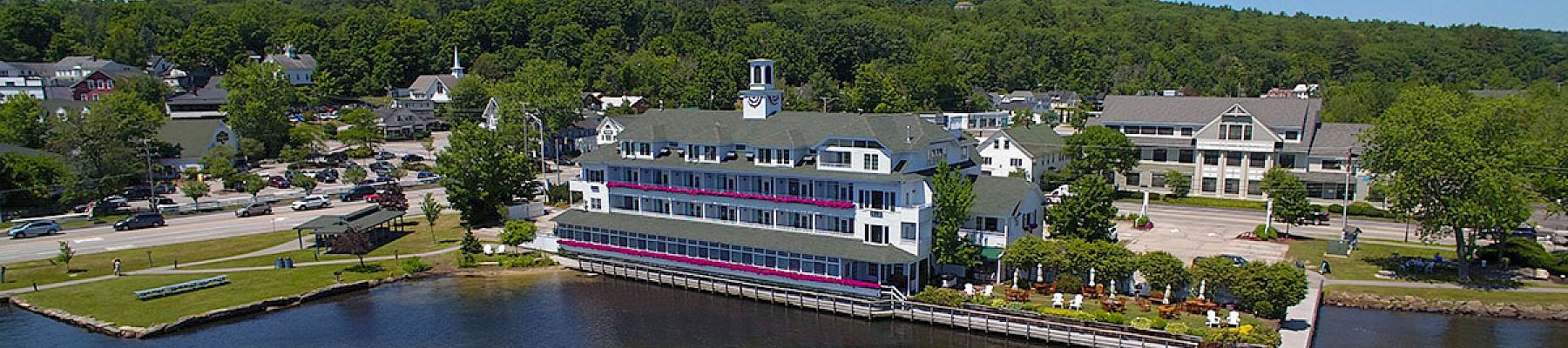 An aerial view of a waterfront area with a multi-story building, a pier, and a boat docked at the pier. Surrounding buildings and trees are visible.