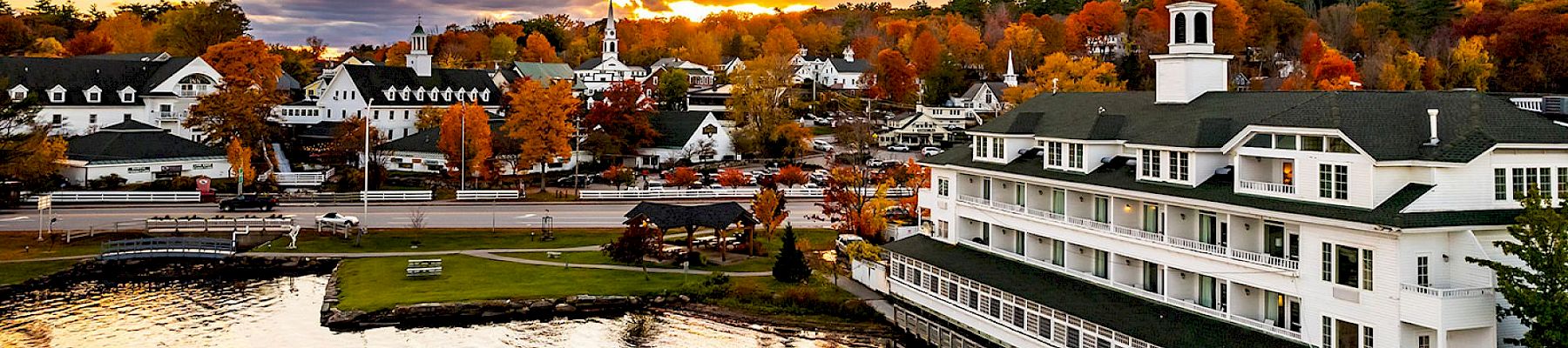 The image shows a picturesque lakeside town during sunset with colorful autumn trees, charming buildings, and serene water reflecting the sky.