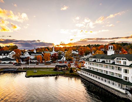 The image shows a picturesque lakeside town during sunset with colorful autumn trees, charming buildings, and serene water reflecting the sky.