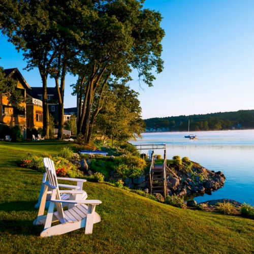 Scenic lakeside view with Adirondack chairs on a grassy shore, houses, trees, a dock, and a boat on the calm water under a clear sky.