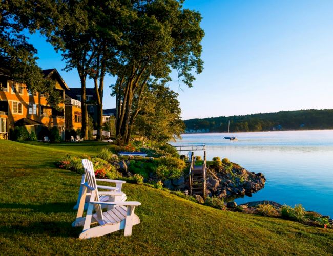 Scenic lakeside view with Adirondack chairs on a grassy shore, houses, trees, a dock, and a boat on the calm water under a clear sky.