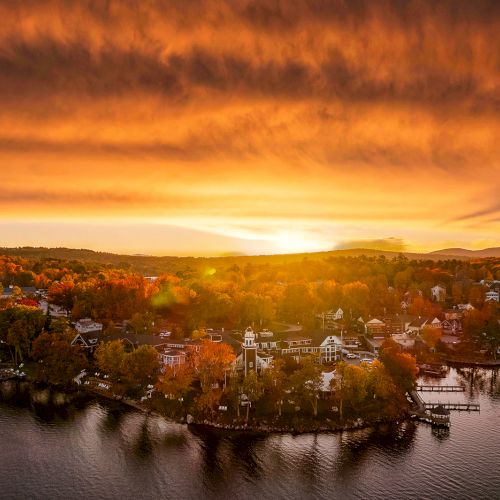 A serene lakeside village is captured at sunset, with a dramatic sky and the warm glow reflecting on the water, enhancing the autumn hues.