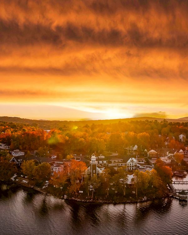 A serene lakeside village is captured at sunset, with a dramatic sky and the warm glow reflecting on the water, enhancing the autumn hues.