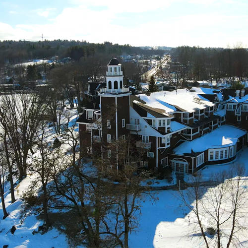 A large, snow-covered building with a tower near a frozen lake, surrounded by trees and a landscape extending into the distance.