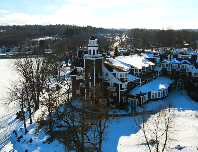 A large, snow-covered building with a tower near a frozen lake, surrounded by trees and a landscape extending into the distance.