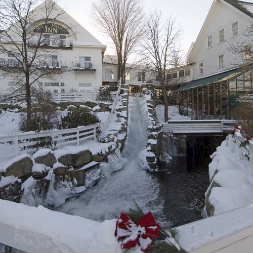 A snowy scene with a frozen stream, decorated bridge, and houses in the background, creating a winter holiday atmosphere.