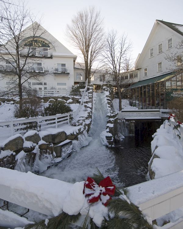 A snowy scene with a frozen stream, decorated bridge, and houses in the background, creating a winter holiday atmosphere.