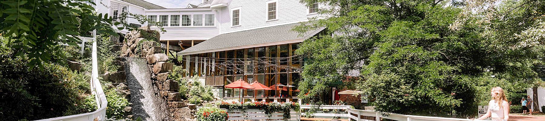 A picturesque building, possibly an inn or restaurant, surrounded by lush greenery, a waterfall, and seating areas with red umbrellas on a sunny day.