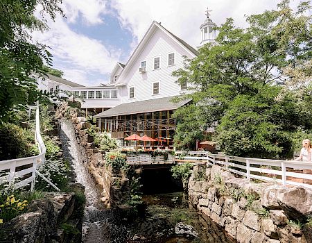 A picturesque building, possibly an inn or restaurant, surrounded by lush greenery, a waterfall, and seating areas with red umbrellas on a sunny day.